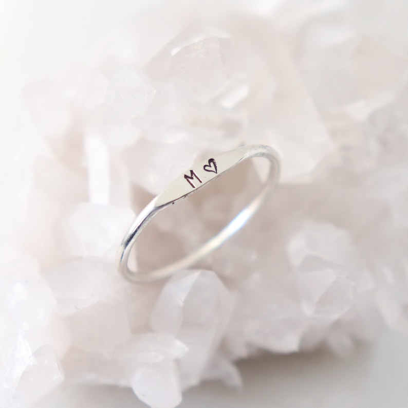 Dainty, sterling silver initial ring with the letter M and a heart stamp sits atop a sparkling white crystal background.  The ring is also available in rose gold filled and gold filled.