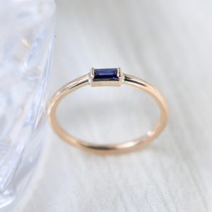 May include: A gold ring with a rectangular, dark blue gemstone. The ring has a thin band and a simple, elegant design. The gemstone is set horizontally in the center of the ring.