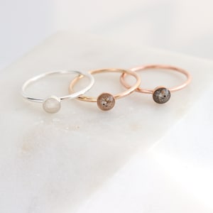 May include: Three thin metal rings with circular settings. The first ring is silver-toned with a smooth white stone. The second and third rings are rose gold-toned, featuring circular settings filled with textured, speckled grey and brown material.
