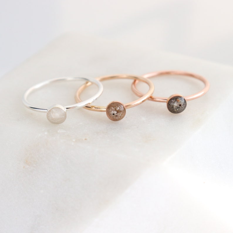 May include: Three thin metal rings with circular settings. The first ring is silver-toned with a smooth white stone. The second and third rings are rose gold-toned, featuring circular settings filled with textured, speckled grey and brown material.