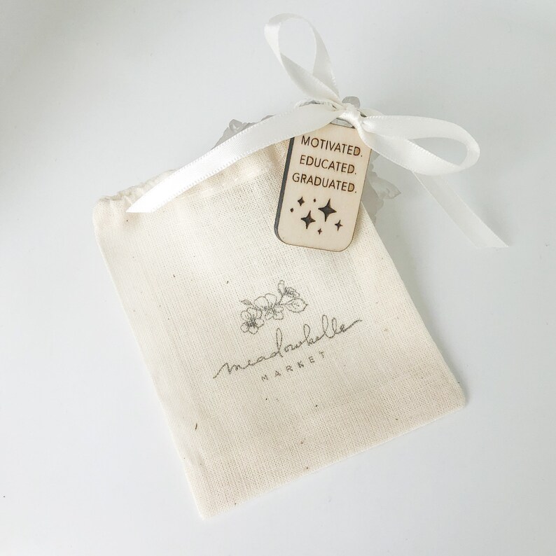 May include: A small, off-white drawstring bag with a floral logo and the text "meadowbelle MARKET" in gray. A wooden tag with the words "MOTIVATED. EDUCATED. GRADUATED." and star graphics is tied to the bag with a white ribbon.