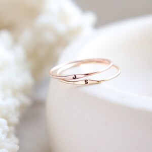 Two slim initial ring bands are stacked on the edge of a jesmonite bowl. The soft, light colored textured cream background is blurred to achieve tight focus on the rose gold J letter ring and gold letter S ring.