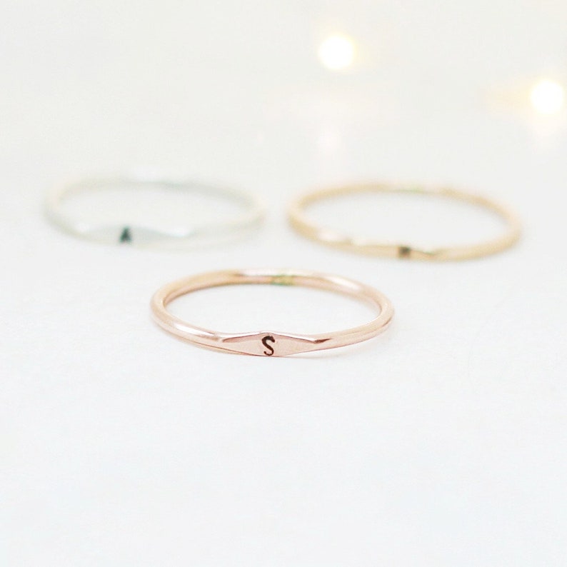 Handmade 14k rose gold slim stackable letter S ring in front of a set of three metal rings placed in a triangle formation on light background, also featuring silver and gold.