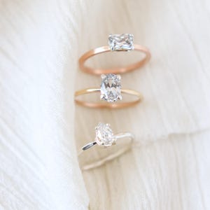 May include: Three engagement rings with different gemstone cuts. The top ring has a rectangular stone on a rose gold band. The middle ring features an oval stone on a gold band. The bottom ring has a pear-shaped stone on a silver band.
