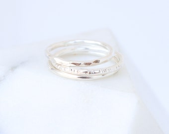 May include: Set of three silver stacking rings. The rings are thin and delicate, with different textures. The top ring has a hammered finish, the middle ring has a textured finish, and the bottom ring is smooth.