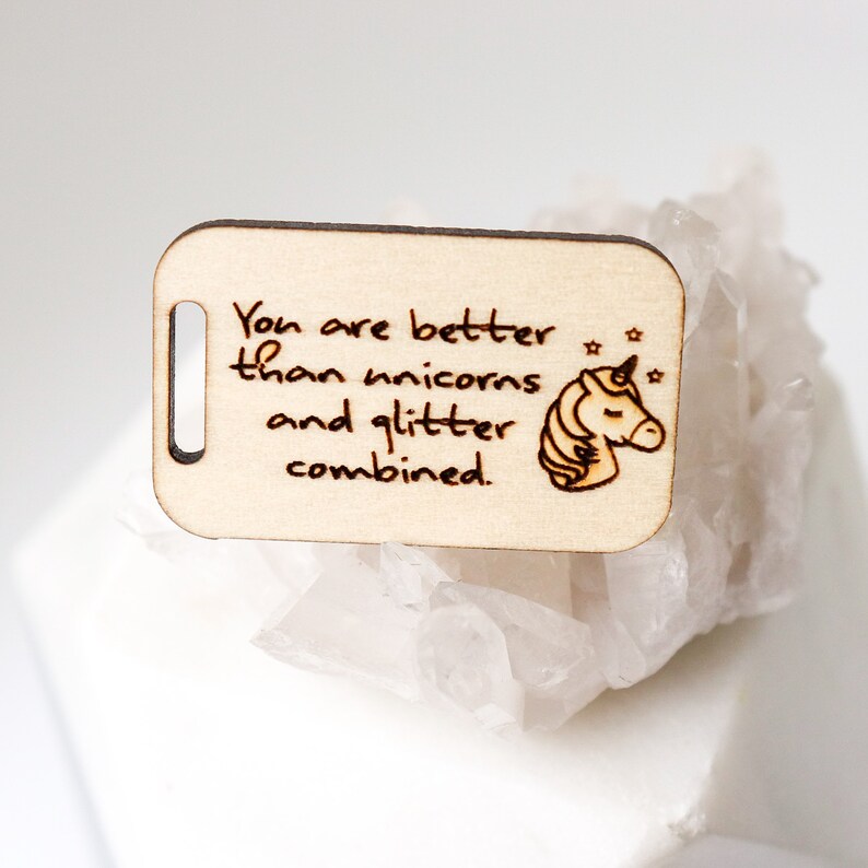May include: A light-colored wooden tag with a cutout hole and rounded corners. The tag features the text "You are better than unicorns and glitter combined." A unicorn head and stars are also etched into the wood.