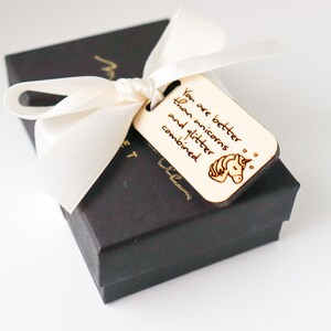 May include: A black gift box tied with a cream-colored ribbon and a wooden tag. The tag is engraved with the words "You are better than unicorns and glitter combined" and a unicorn illustration.