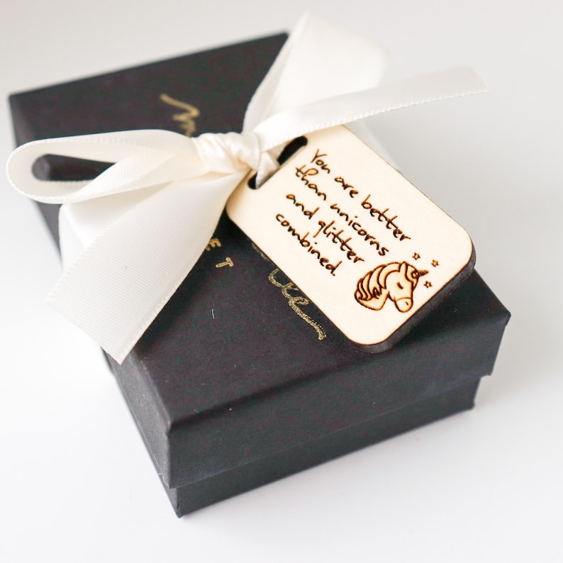 May include: A black gift box tied with a cream-colored ribbon and a wooden tag. The tag is engraved with the words "You are better than unicorns and glitter combined" and a unicorn illustration.