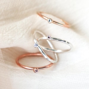 May include: A collection of delicate rings in rose gold and silver. Each ring features a small, circular charm with a single initial or symbol. The rings are displayed on a soft, white fabric, creating a minimalist aesthetic.