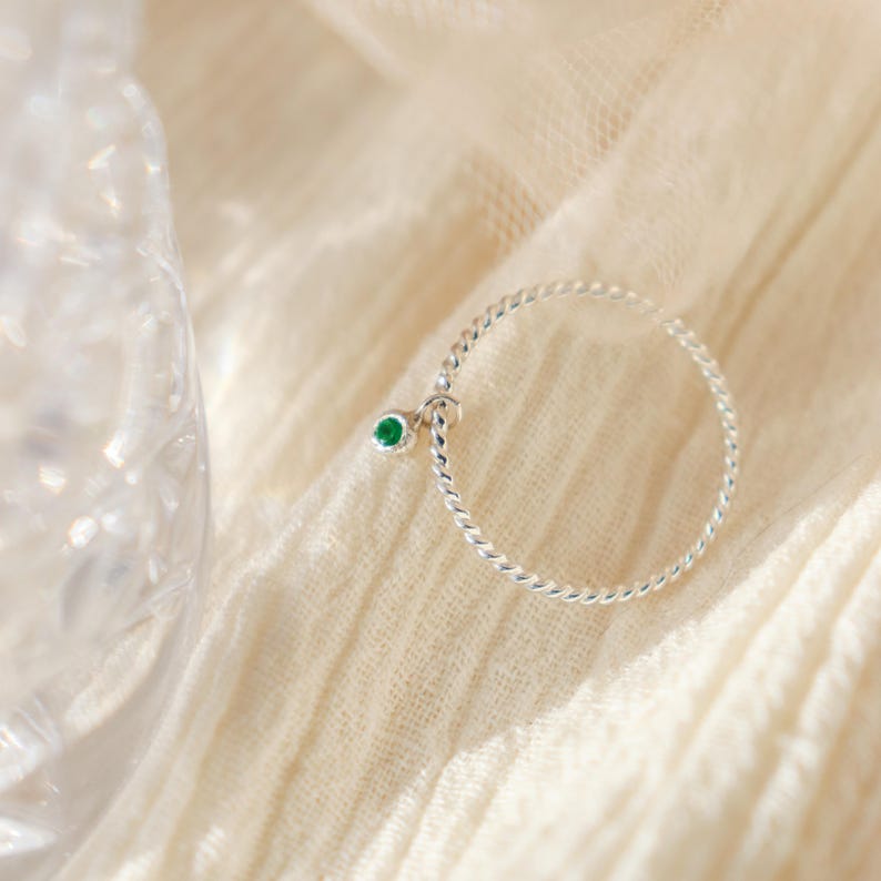K&ouml;nnte beinhalten: Ein filigraner Silberring mit gedrehtem Band. Ein kleiner, runder, gr&uuml;ner Edelstein h&auml;ngt an dem Ring. Der Ring wird auf einem weichen, cremefarbenen Stoff pr&auml;sentiert, mit einem verschwommenen Glasobjekt im Hintergrund.