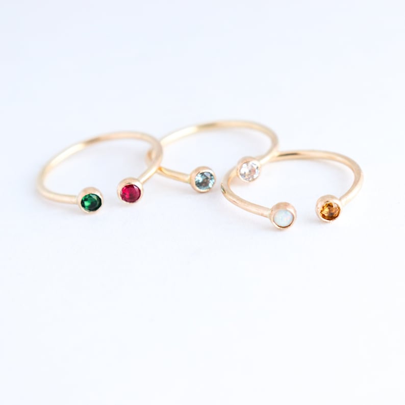 Three gold cuff rings are seen from a top view lying next to each other with glistening gemstones of various colours in each end. Rings are lying on an off-white background.