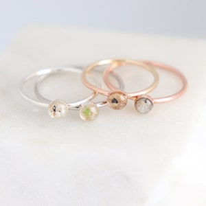 May include: Three delicate rings with small round clear resin insets containing different natural elements. The rings are in silver, gold, and rose gold.