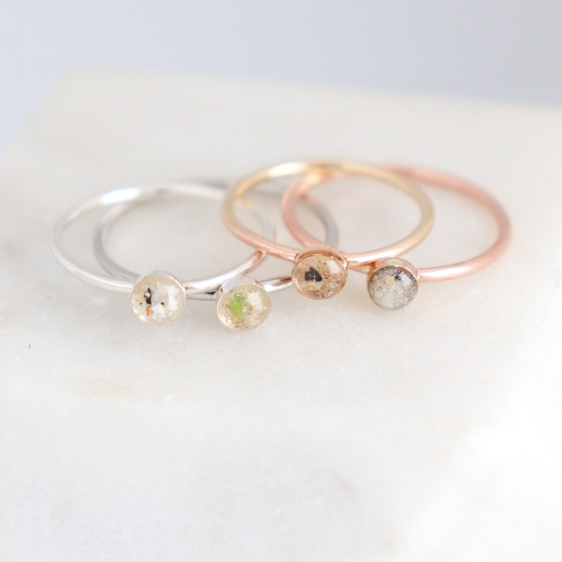 May include: Four thin metal rings featuring small circular settings. Two rings are silver-toned with white and light green speckled inlays. Two rings are rose gold-toned with tan, brown, and grey speckled inlays in the circular center pieces.