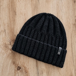 Puede incluir: Un gorro de punto negro sólido que presenta una textura acanalada gruesa en toda la corona y un borde ancho doblado. Una pequeña etiqueta rectangular de cuero sintético gris oscuro está unida al lado derecho del borde con un remache metálico plateado.