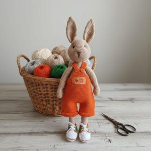 Puede incluir: Un conejo hecho a mano de ganchillo con un mono naranja y zapatillas blancas con detalles verdes. El conejo tiene orejas largas y un pequeño parche que dice "WITH LOVE". Una cesta de ovillos de lana está en el fondo.
