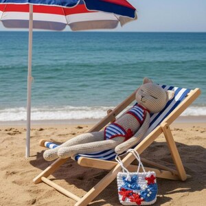 May include: A crocheted cat wearing a red, white, and blue striped swimsuit is lounging on a blue and white striped beach chair. A matching beach bag with red and blue starfish and seashell designs sits nearby, under a red, white, and blue striped umbrella on a sandy beach.