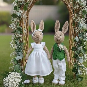 Könnte beinhalten: Zwei gehäkelte Hasenpuppen, gekleidet für eine Hochzeit. Einer trägt ein weißes Kleid und ein Stirnband, der andere eine grüne Weste und eine weiße Hose. Sie stehen vor einem Blumenbogen auf einer Wiese, eine bezaubernde Szene.