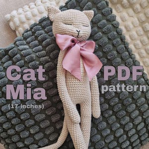 Puede incluir: Muñeca de gato de ganchillo beige, de unos 43 cm de altura, con lazo rosa y ojos cerrados. El texto "Cat Mia" y "PDF pattern" son visibles. El gato descansa sobre un cojín verde texturizado.
