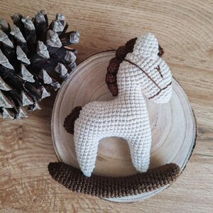 May include: A handmade, crocheted rocking horse toy in cream and brown. The horse features a cream-colored body, brown mane, tail, and rocker base. The toy is placed on a wooden slice, with a pine cone in the background.