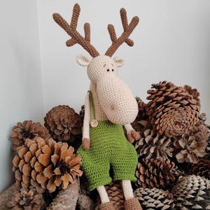 Puede incluir: Muñeco de alce de ganchillo hecho a mano con cuernos marrones, cuerpo beige y peto verde. El muñeco tiene una nariz grande y está sentado sobre un lecho de piñas. Un artículo artesanal caprichoso.