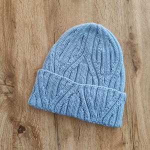 Pode incluir: Um gorro de malha azul-claro com um padrão de tranças verticais em todo o corpo e uma aba dobrada. O gorro é feito de um material de fio macio com uma textura consistente e uma cor azul pastel uniforme em toda a peça.