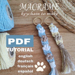 May include: A PDF tutorial for making a macrame keychain with paw print designs. The tutorial is available in English, German, French, and Spanish. The image shows two completed keychains, one in blue and white and the other in yellow and white.