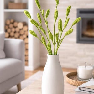 May include: A white ceramic vase holds a bouquet of green crochet stems with textured buds. The vase sits on a light wood table, with a neutral-toned living room in the background. The stems add a touch of handmade charm to the decor.