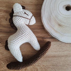 May include: A handmade crochet rocking horse toy in cream and brown. The horse features a cream-colored body, brown mane and rocker base, and brown facial details. The toy is designed for young children.