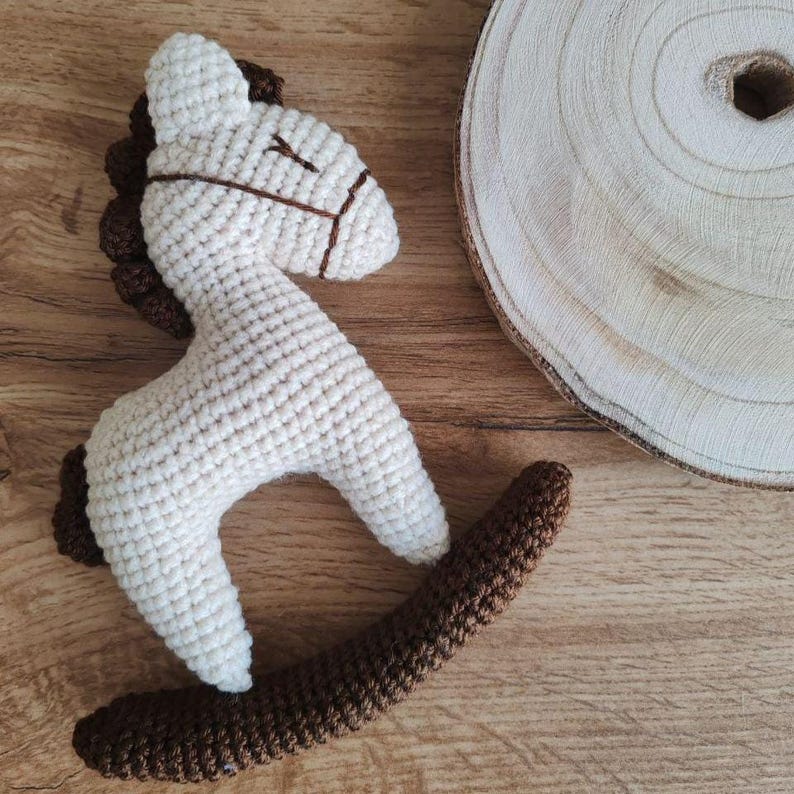 May include: A handmade crochet rocking horse toy in cream and brown. The horse features a cream-colored body, brown mane and rocker base, and brown facial details. The toy is designed for young children.