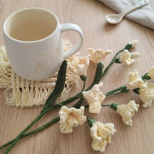 May include: A white ceramic mug with a bird and floral design, resting on a cream-colored macrame coaster. Crochet flowers with green stems and pale yellow petals are arranged nearby, along with a small ceramic spoon.