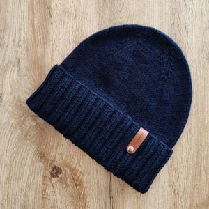 May include: A navy blue knit beanie hat with a folded brim. A small brown leather tag with the words "HAND MADE" is attached to the brim. The hat is made of a thick, textured yarn.