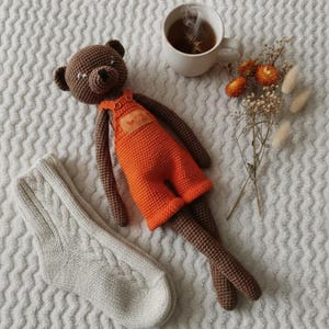 Könnte beinhalten: Ein handgefertigter brauner Häkelbär mit orangefarbenen Latzhosen und einem herzförmigen Aufnäher. Ein Paar Zopfstricksocken und eine Tasse Tee mit Blumen sind ebenfalls im Bild. Der Bär liegt auf einer weißen, strukturierten Oberfläche.