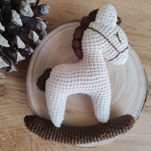 May include: A handmade crochet rocking horse in cream and brown. The toy horse features a cream-colored body, brown mane, tail, and rocker base. The horse is placed on a wooden slice, with a pine cone in the background.
