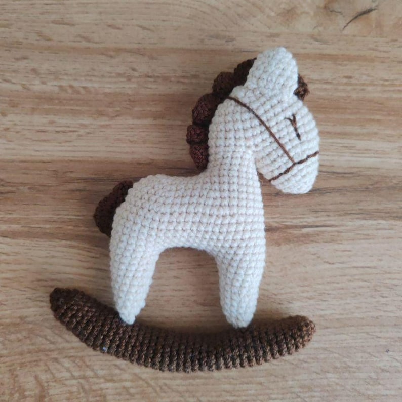May include: A crocheted rocking horse toy in cream and brown. The horse has a cream-colored body, a brown mane and tail, and a brown bridle. The toy is designed to sit on a brown rocker base.