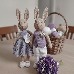 May include: Two crocheted bunny dolls, one in a lavender dress with a flower and the other in a purple vest and white pants. A bouquet of purple flowers and a basket of yarn balls are in the background. The bunnies are wearing white shoes.