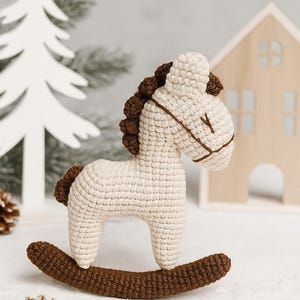 Puede incluir: Un caballo balancín de ganchillo en crema y marrón. El caballo tiene un cuerpo color crema, una melena y cola marrones, y se asienta sobre una base balancín marrón. El fondo incluye un árbol de Navidad blanco y una pequeña casa de madera.