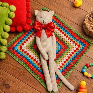 May include: A beige cat-shaped stuffed animal with a red bow tie, lying on a colorful crocheted blanket. The blanket has a green border and a vibrant pattern of red, orange, blue, and white squares. A yellow rubber duck and other toys are nearby.