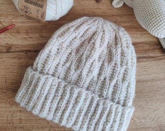 May include: A cream-colored, hand-knitted beanie hat with a ribbed brim and a textured, cable-knit design. The hat is made of soft yarn and is suitable for cold weather. A ball of yarn and a crochet animal are in the background.