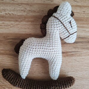 May include: A crocheted rocking horse toy in cream and brown. The horse has a cream-colored body, a brown mane and tail, and a brown rocker base. The horse's face has brown details for the eyes and bridle. The toy is made of soft yarn.