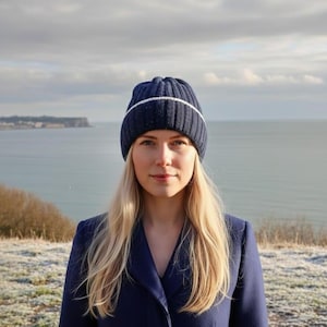 May include: A navy blue knit beanie with a white stripe is worn. The person is wearing a navy blue coat. The background shows a body of water and a cloudy sky.