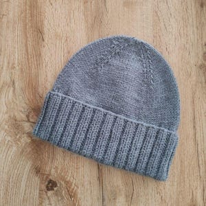 Może przedstawiać: Szara dzianinowa czapka z wywiniętym ściągaczem. Czapka wykonana jest z miękkiej, teksturowanej przędzy i ma za zadanie utrzymywać ciepło głowy. Czapka jest pokazana na drewnianej powierzchni.