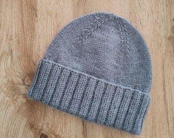 May include: A gray knitted beanie hat with a ribbed brim. The hat is made of a soft, textured yarn and is designed to keep the head warm. The hat is shown on a wooden surface.