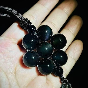 May include: A black beaded necklace featuring a cluster of seven large, round, polished obsidian beads. The beads are strung together with a black cord and have a glossy finish. The necklace is held in a hand against a dark background.