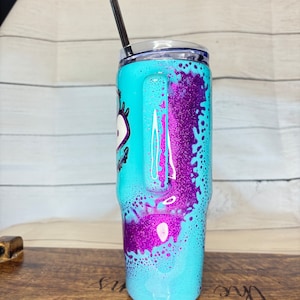 Puede incluir: Un vaso turquesa con asa, con un dise&ntilde;o de purpurina morada y un gr&aacute;fico de ojo de dibujos animados. Incluye una tapa transparente y una pajita negra. El dise&ntilde;o se aplica al exterior del vaso de acero inoxidable.