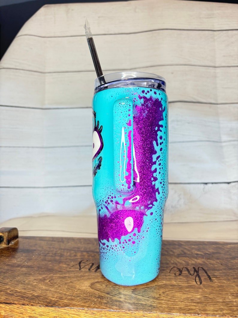 Puede incluir: Un vaso turquesa con asa, con un dise&ntilde;o de purpurina morada y un gr&aacute;fico de ojo de dibujos animados. Incluye una tapa transparente y una pajita negra. El dise&ntilde;o se aplica al exterior del vaso de acero inoxidable.