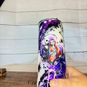 May include: A stainless steel tumbler featuring a design of Jack Skellington and Sally from The Nightmare Before Christmas. The tumbler has a purple, black, and white glitter design. The characters are in a heart-shaped frame.