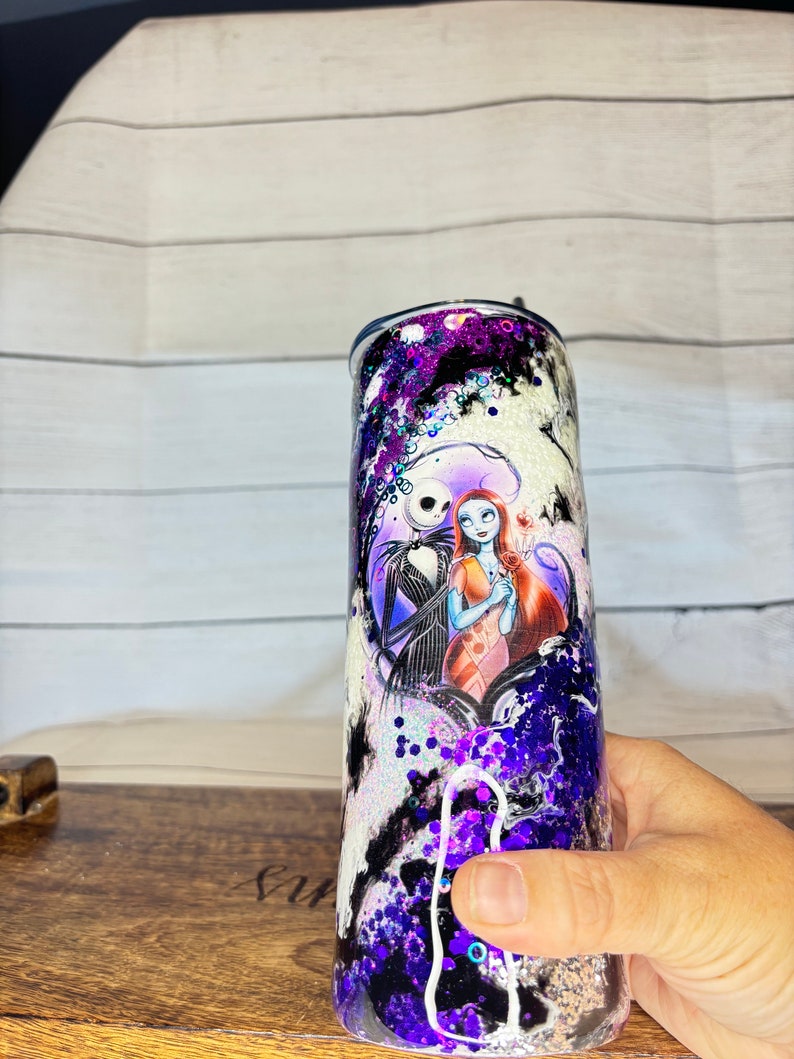 May include: A stainless steel tumbler featuring a design of Jack Skellington and Sally from The Nightmare Before Christmas. The tumbler has a purple, black, and white glitter design. The characters are in a heart-shaped frame.