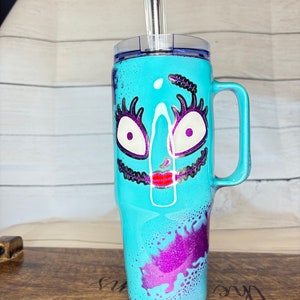 Puede incluir: Un vaso de color turquesa con asa, con un dise&ntilde;o de cara de dibujos animados con ojos blancos, pesta&ntilde;as moradas y boca roja. El vaso tiene tapa transparente y pajita de acero inoxidable. Acentos de purpurina morada.