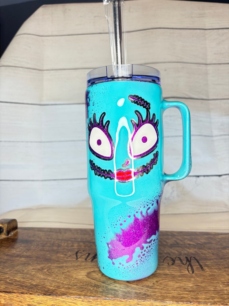 Puede incluir: Un vaso de color turquesa con asa, con un dise&ntilde;o de cara de dibujos animados con ojos blancos, pesta&ntilde;as moradas y boca roja. El vaso tiene tapa transparente y pajita de acero inoxidable. Acentos de purpurina morada.