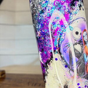 May include: A glitter-filled tumbler featuring a design from "The Nightmare Before Christmas." The tumbler has a black lid and is filled with purple, blue, and silver glitter. The design includes Jack Skellington and Sally.